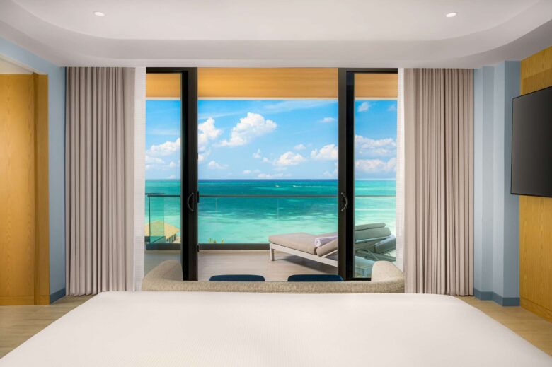 View from a modern hotel room with a bed, open sliding doors, and a balcony overlooking turquoise ocean water and a blue sky with clouds. Lounge chairs are on the balcony.