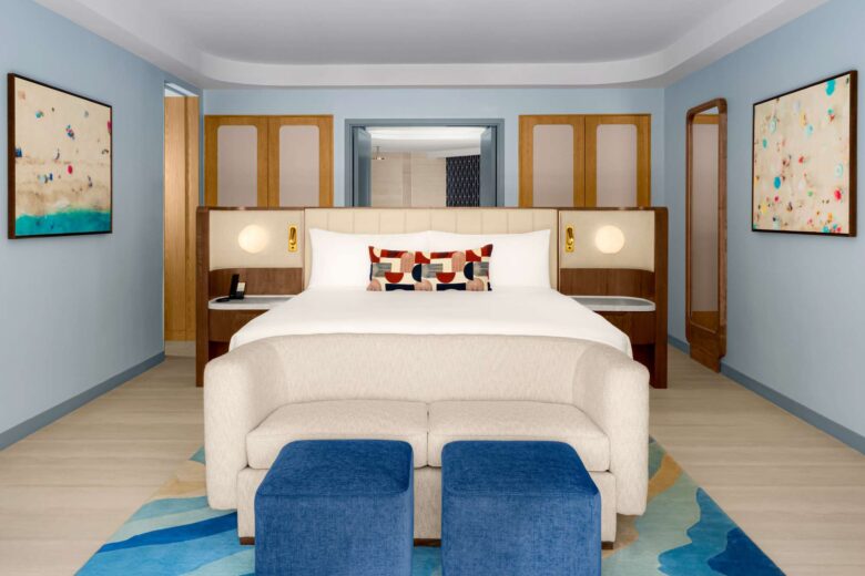 Modern hotel room with a king bed, abstract pillows, a cream sofa, two blue ottomans, wooden accents, and colorful abstract art on light blue walls. Bright, clean, and minimalist decor.