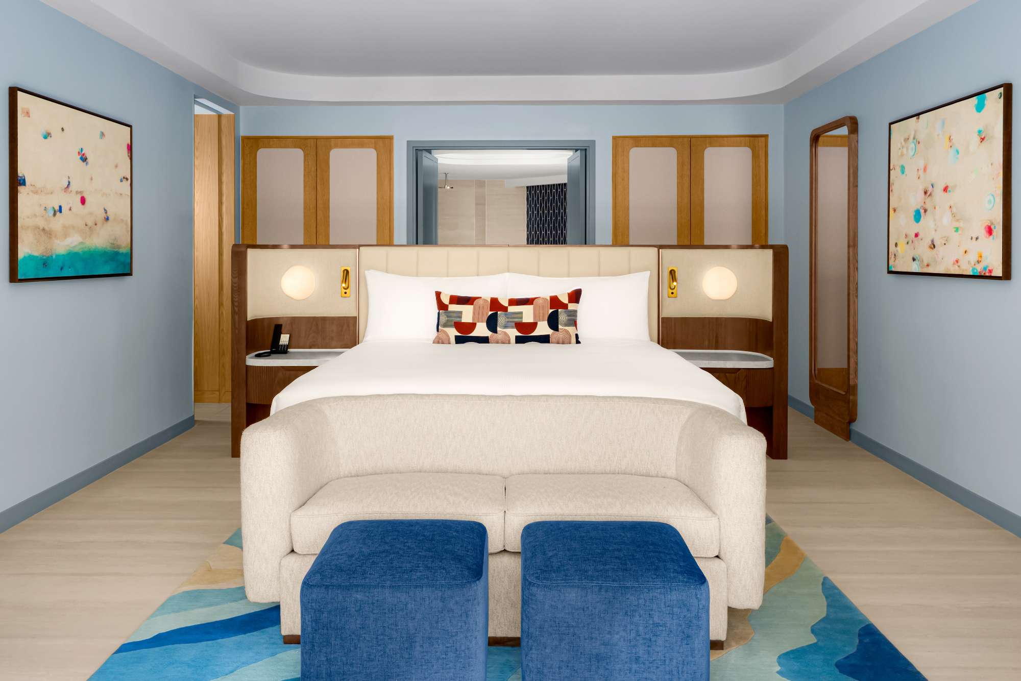Modern hotel room with a king bed, abstract pillows, a cream sofa, two blue ottomans, wooden accents, and colorful abstract art on light blue walls. Bright, clean, and minimalist decor.