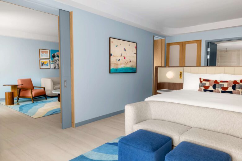 A modern hotel suite with a bed, sofa, blue ottomans, colorful decor, and an adjoining sitting area featuring chairs, a table, and wall art. The room has light blue walls and wood accents.