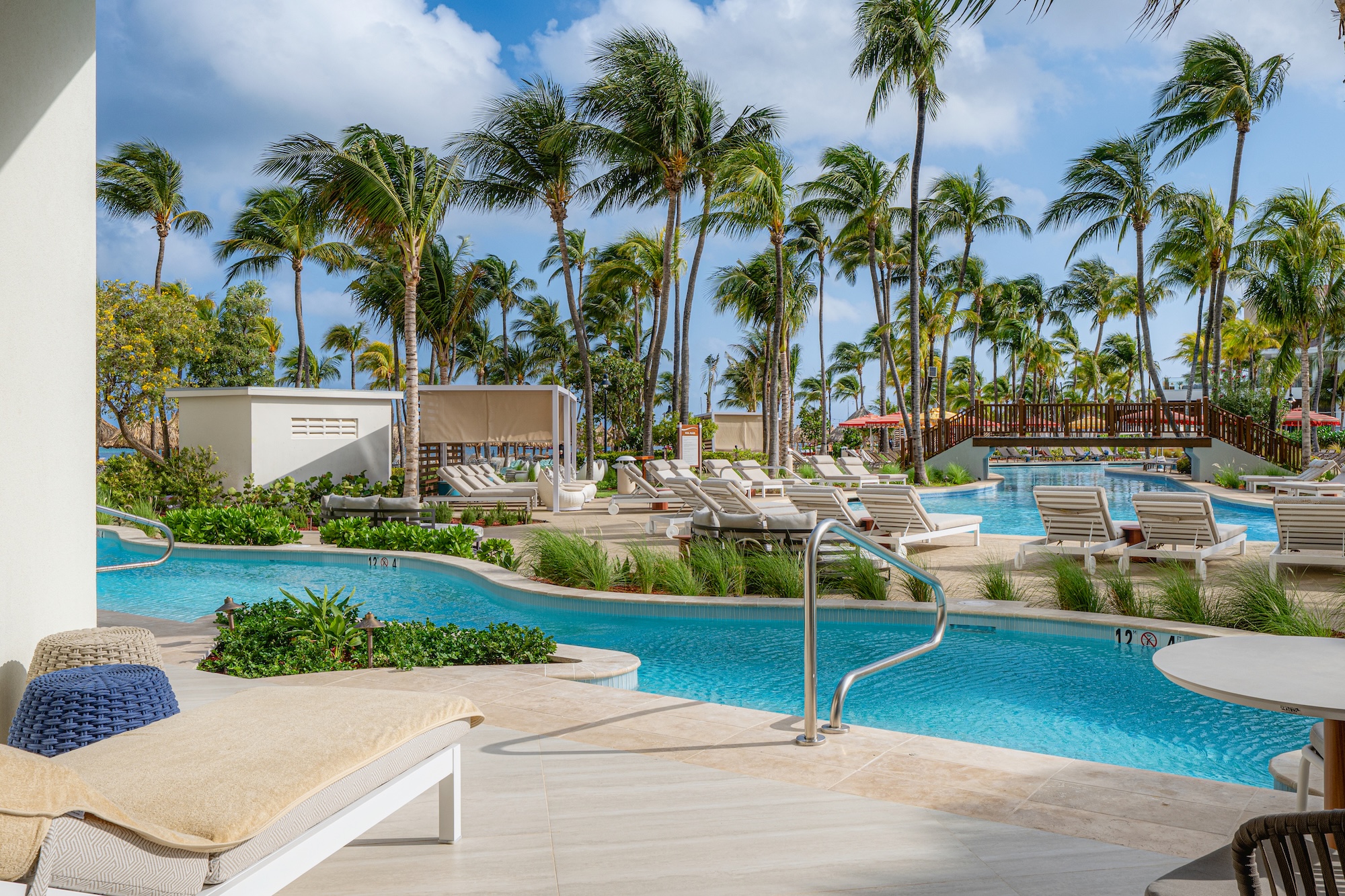 A resort pool area with lounge chairs, palm trees, and a wooden bridge. Blue water winds through the scene, with cabanas and lush greenery under a sunny sky with scattered clouds.