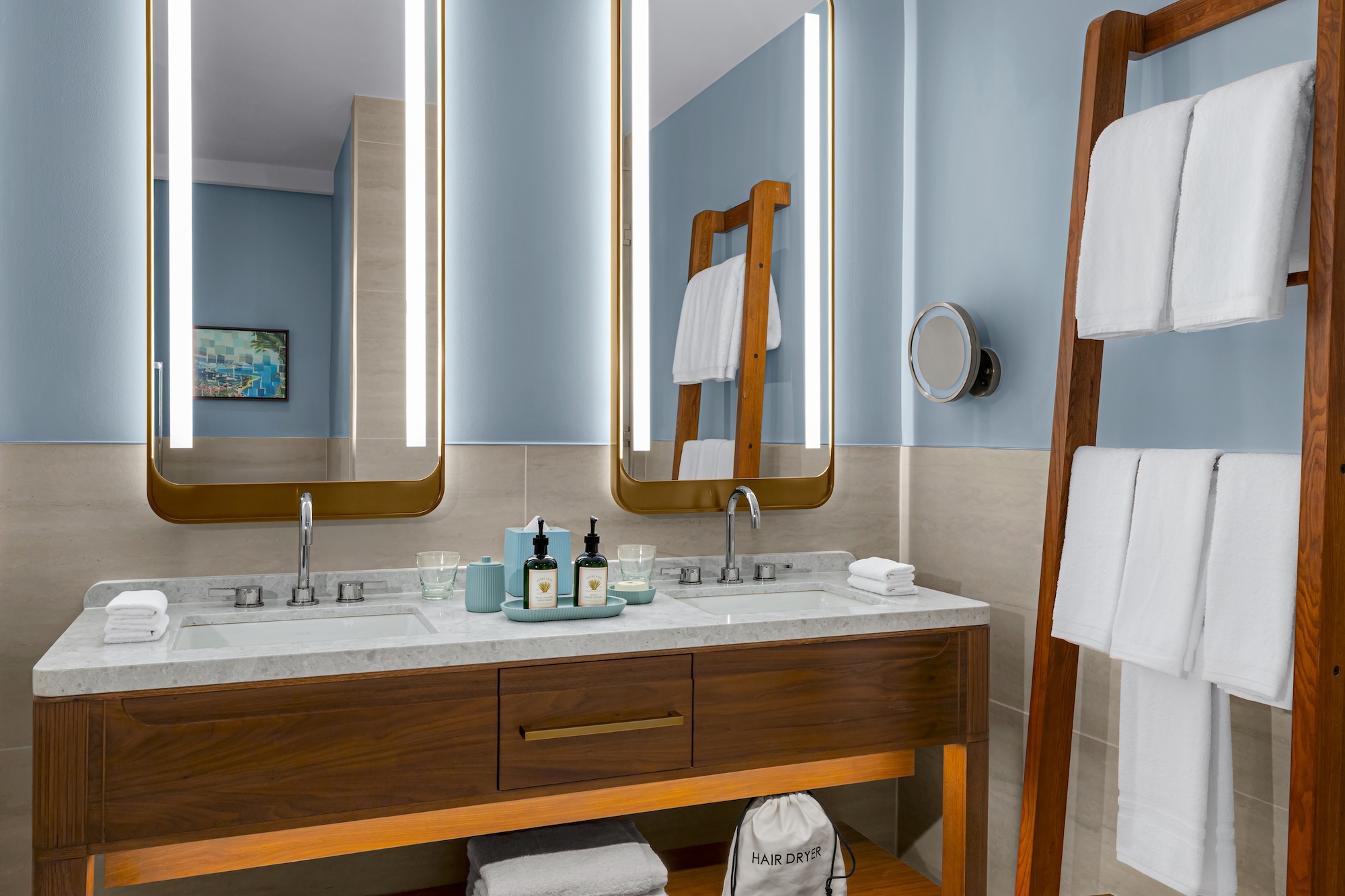 A modern bathroom with a double sink vanity, two illuminated mirrors, toiletries, and rolled towels. A wooden ladder holds white towels beside the vanity. The walls are light blue.