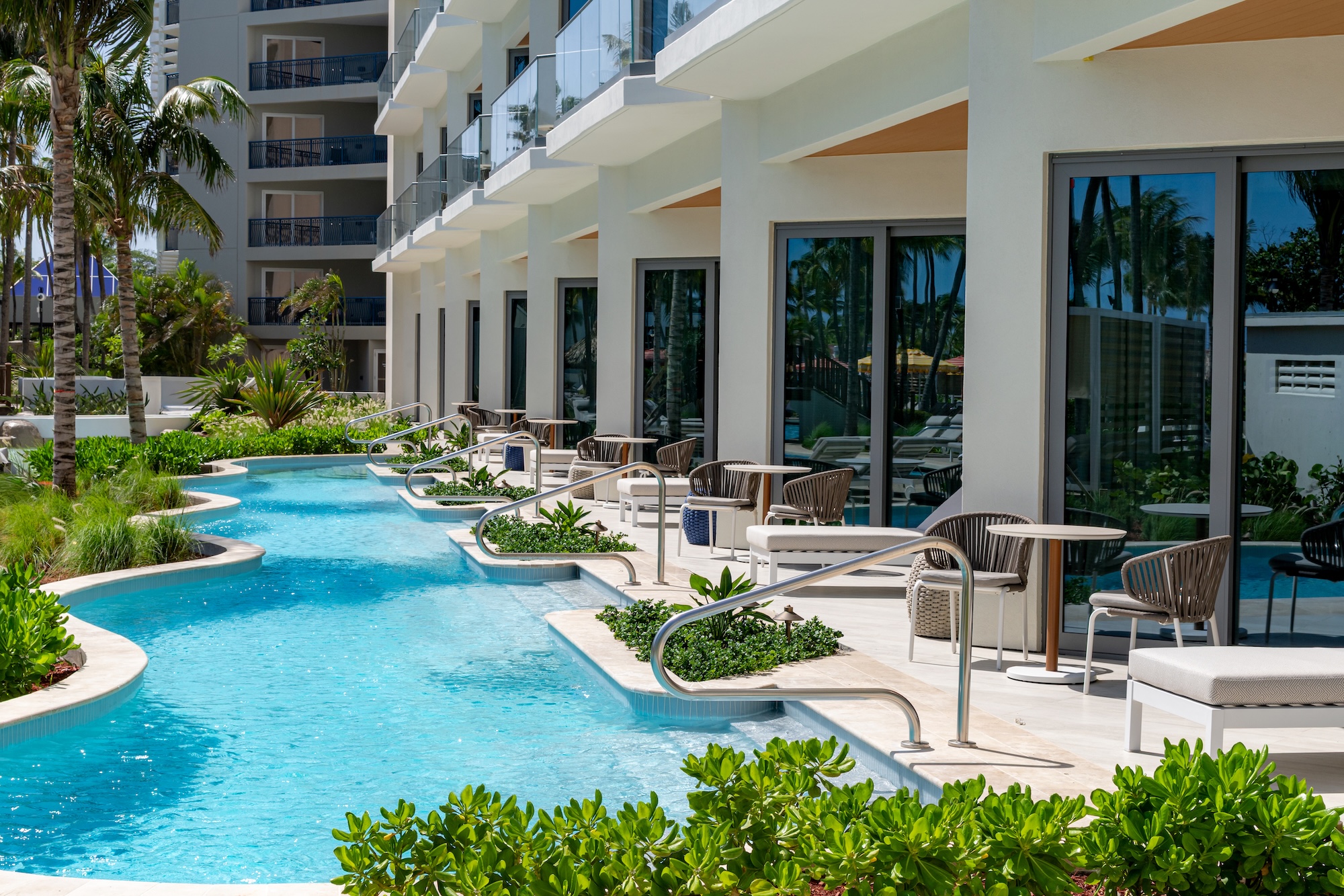 A curved pool runs alongside a modern hotel, with lounge chairs and small tables on private patios. Lush plants border the pool, and palm trees are reflected in large glass windows.