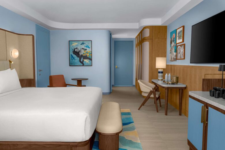 Modern hotel room with a neatly made bed, wooden furniture, artwork on blue walls, a flat-screen TV, desk with lamp, and a minibar setup beside a small table and chair.