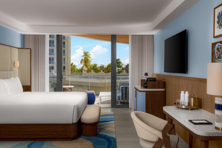 Modern hotel room with a large bed, TV, desk, and coffee maker. Floor-to-ceiling windows open to a balcony with palm trees and buildings visible outside. Light blue walls and contemporary decor.