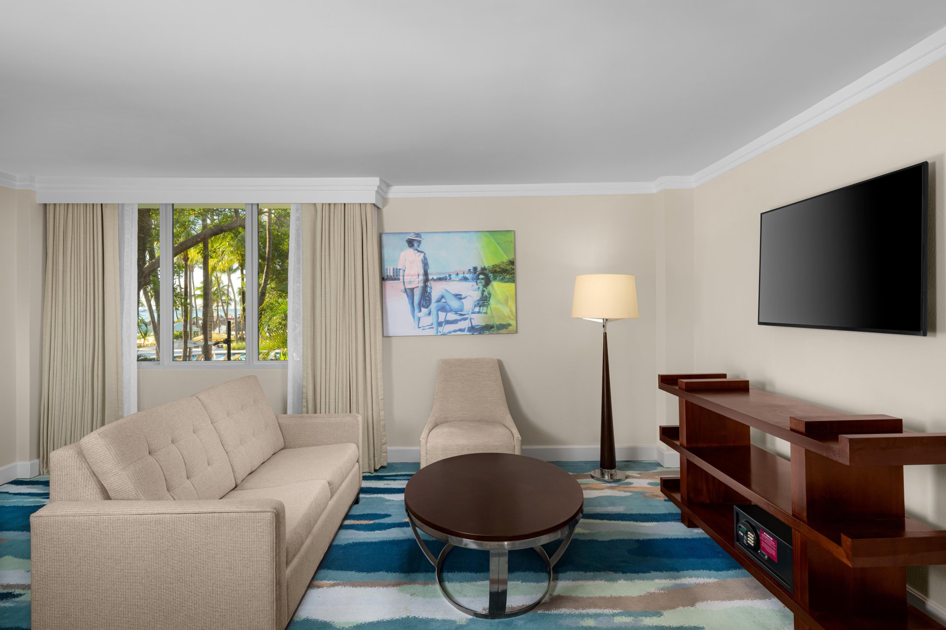 A hotel living room with a beige sofa, a matching chaise lounge, a round wooden coffee table, a floor lamp, a wall-mounted TV, abstract art, and large window with trees visible outside.