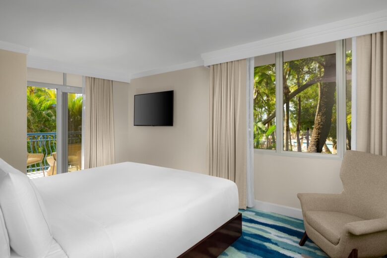 A hotel room with a large bed, wall-mounted TV, armchair, and floor-to-ceiling windows showing tropical trees and a balcony outside. The room has light-colored walls and a blue-patterned carpet.