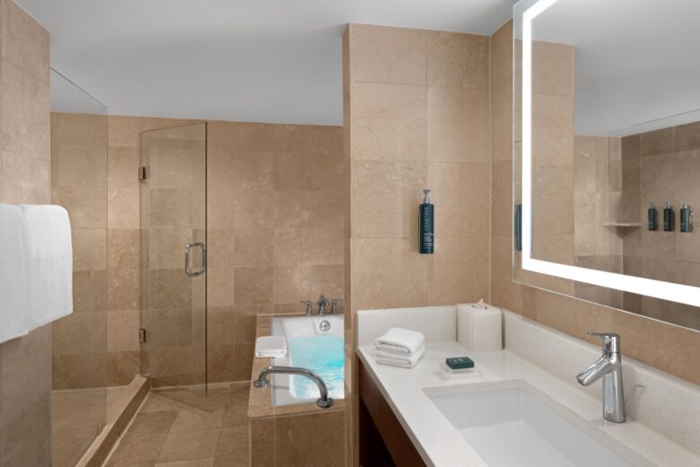 Modern bathroom with beige marble walls, a bathtub filled with water, glass-enclosed shower, large illuminated mirror above a white sink, and neatly folded towels on the countertop.