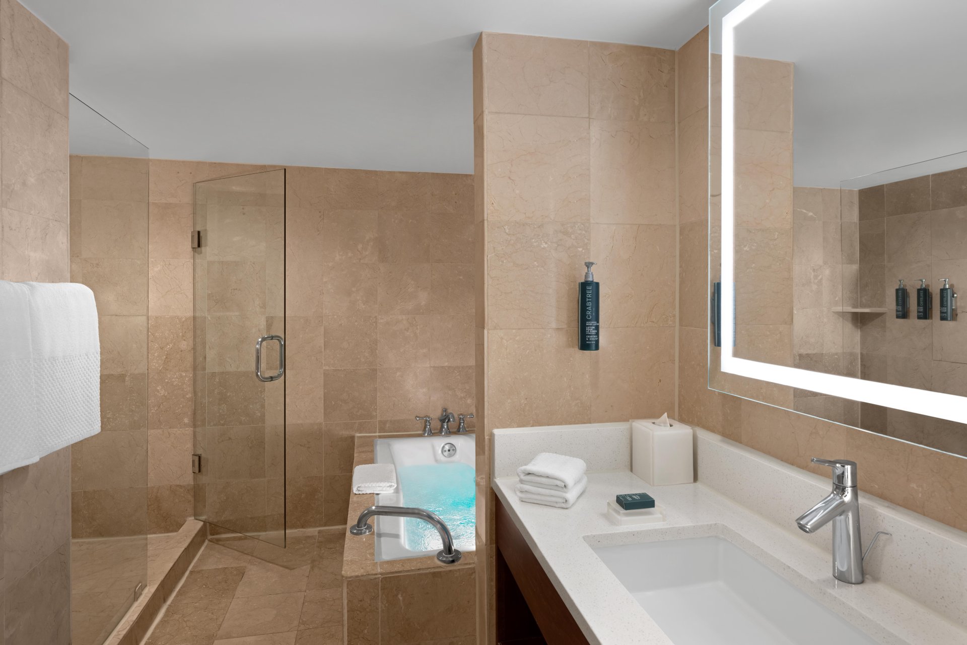 Modern bathroom with beige marble walls, a bathtub filled with water, glass-enclosed shower, large illuminated mirror above a white sink, and neatly folded towels on the countertop.