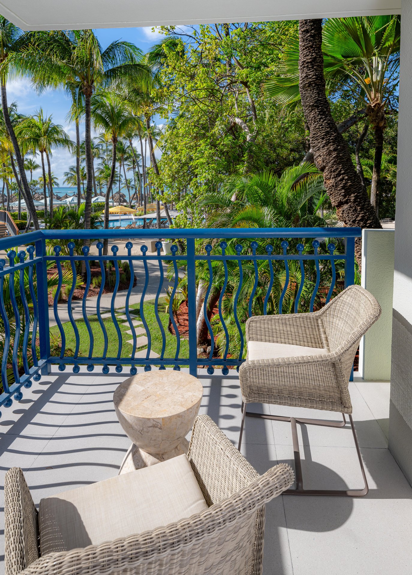 A balcony with two wicker chairs and a small stone table overlooks a lush, tropical garden with palm trees and a path leading toward the beach under a sunny sky.