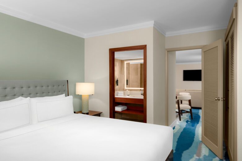 A hotel room with a neatly-made white bed, a lamp on a nightstand, an open door to a bathroom with towels, and another open door leading to a sitting area with chairs and a television.