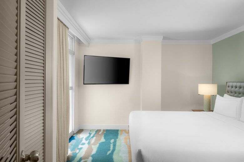 A hotel room with a large bed, a pastel green accent wall, a bedside table with a lamp, a wall-mounted TV, patterned carpet, and curtains covering a window or balcony door.