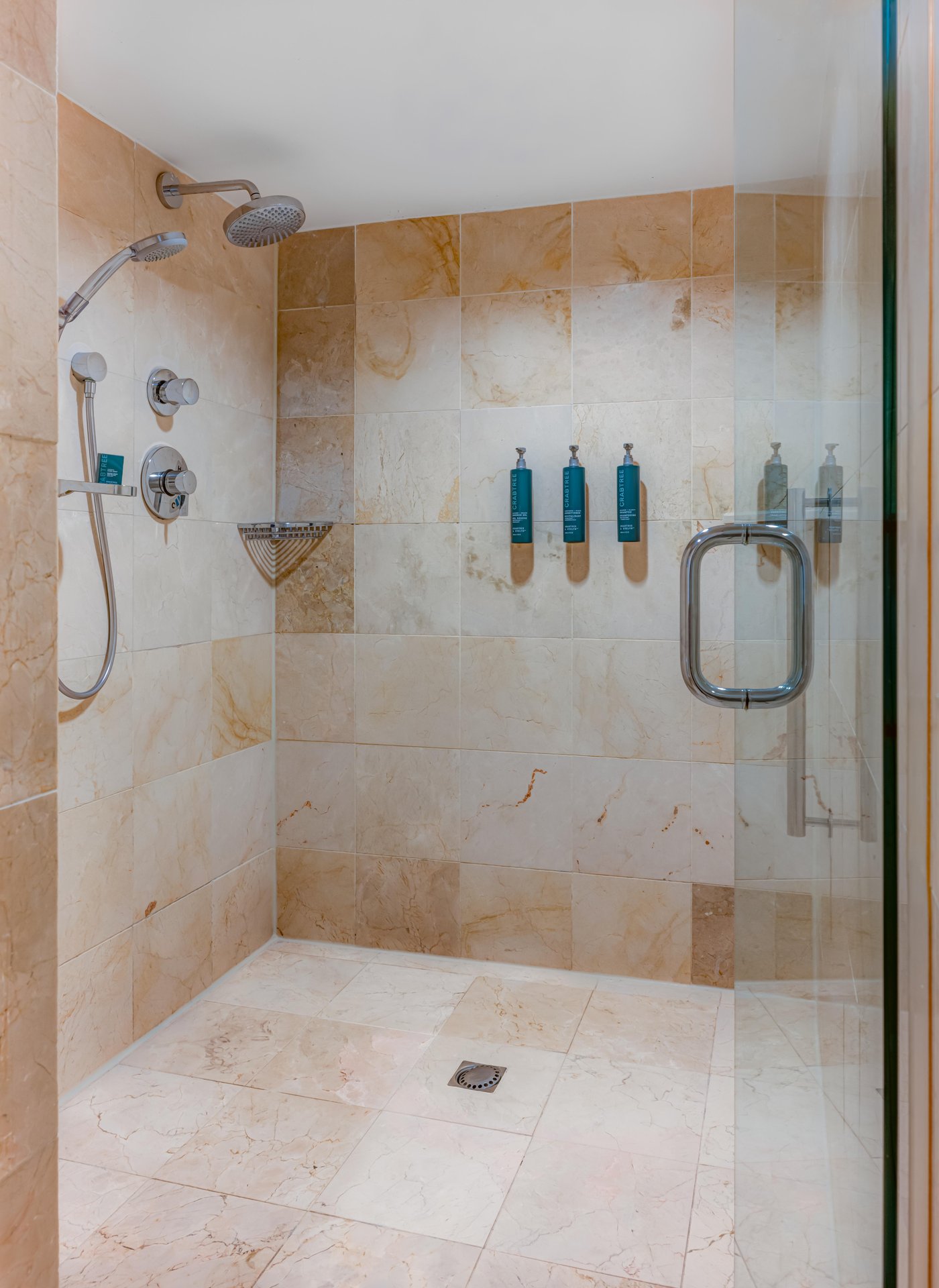 A modern tiled shower with a glass door, dual showerheads, built-in controls, wall-mounted soap dispensers, and a small metal corner shelf. The walls and floor are covered in beige marble tiles.