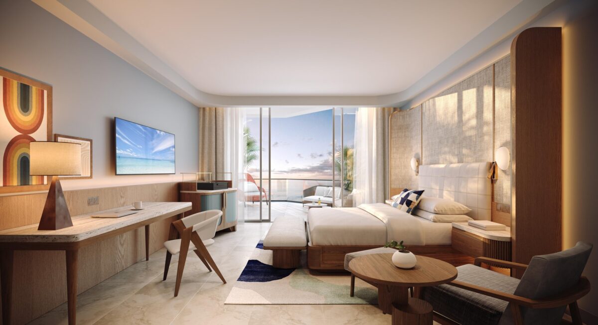 Modern hotel room with a large bed, desk, TV, and seating area. Glass doors open to a balcony with ocean views, letting in natural light. Decor is neutral with wood accents and colorful wall art.