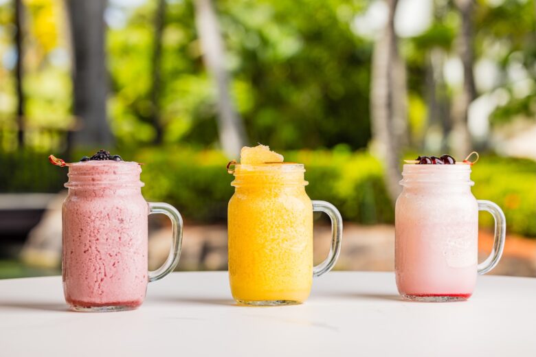 Three mason jar glasses filled with colorful smoothies—pink with berries, yellow with a frosty texture, and light pink with cherries—sit on a white table outdoors, with greenery blurred in the background.
