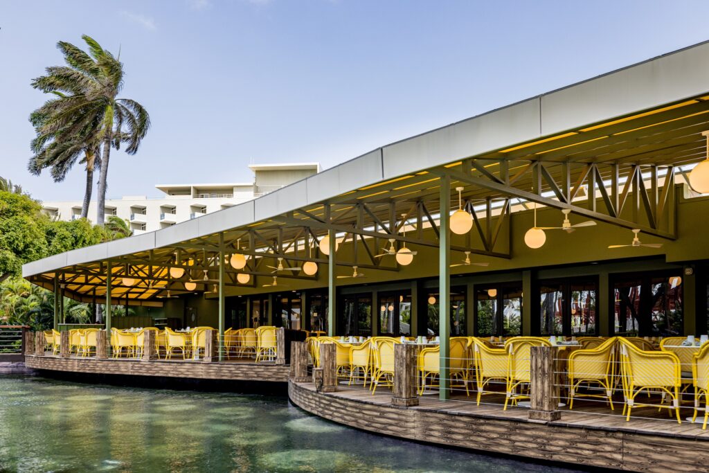 Outdoor restaurant with yellow chairs and round hanging lights is set on a wooden deck over water, surrounded by palm trees and greenery, under a clear blue sky.