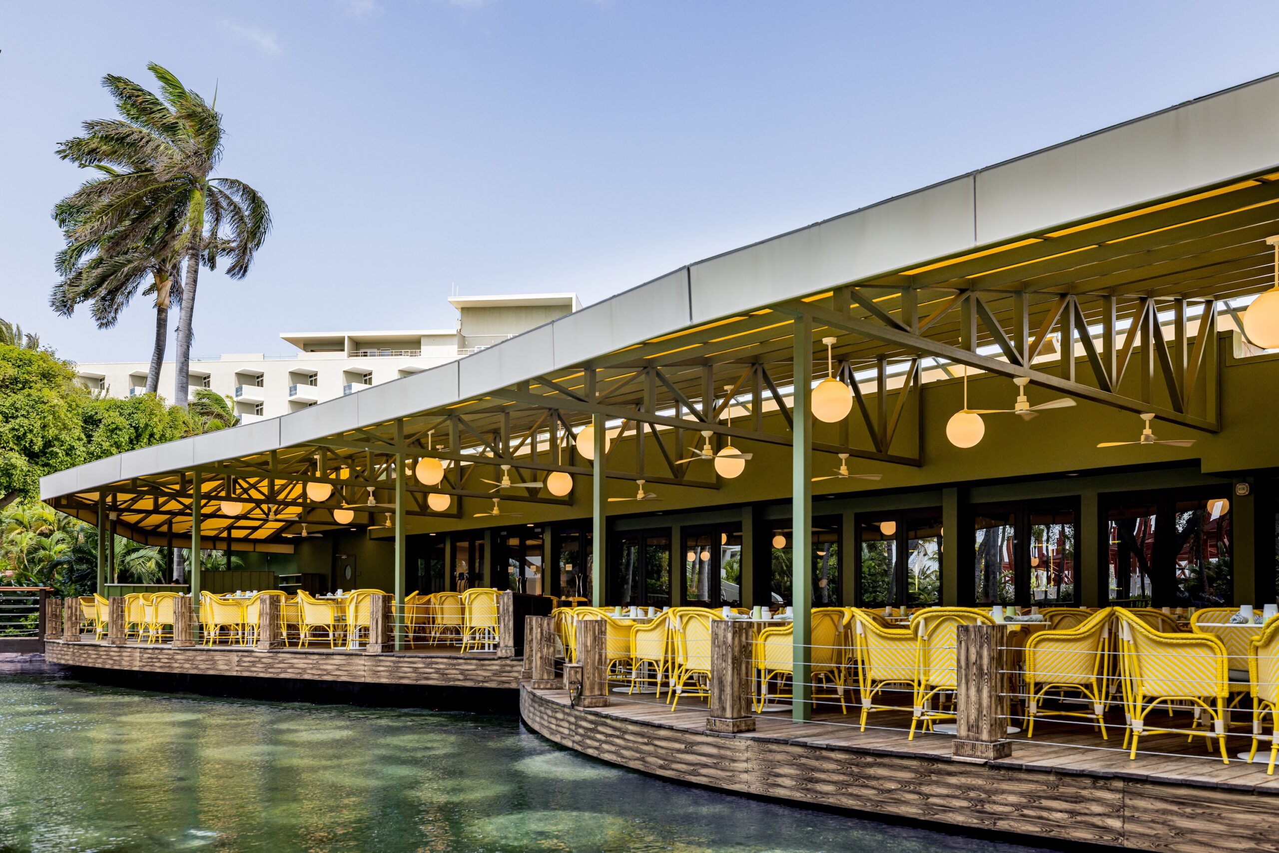Outdoor restaurant with yellow chairs and round hanging lights is set on a wooden deck over water, surrounded by palm trees and greenery, under a clear blue sky.