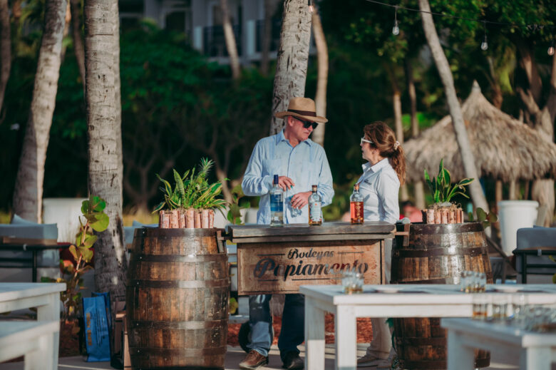 A man and woman stand behind a rustic outdoor bar made of wooden barrels and a wooden plank labeled "Bodegas Pepiamonte," surrounded by palm trees and greenery.
