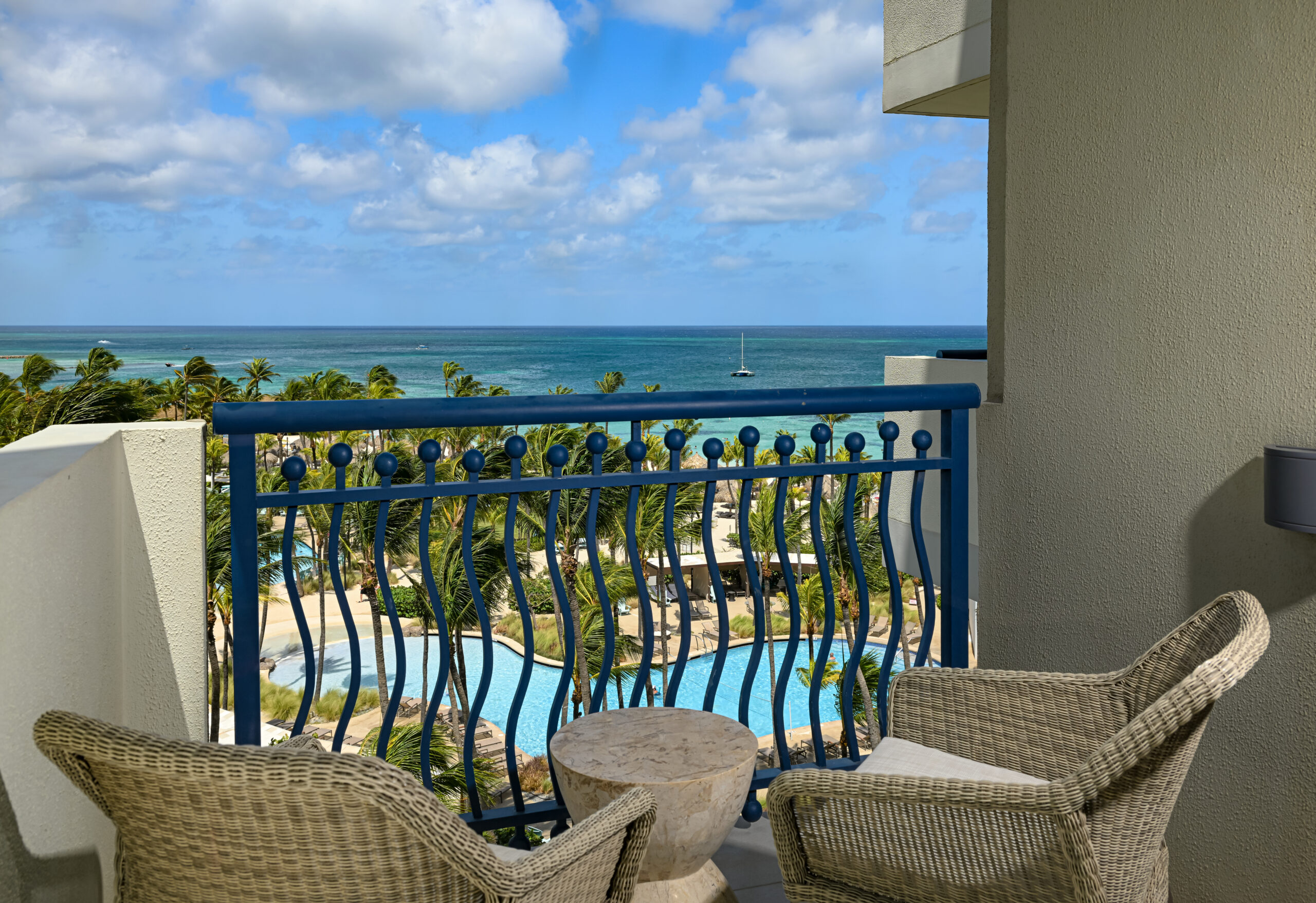 A balcony with two wicker chairs and a small round table overlooks a pool, palm trees, and the ocean with a sailboat in the distance under a partly cloudy blue sky.
