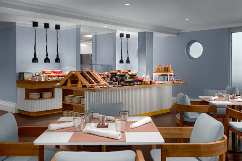 A modern buffet restaurant with a central serving station offering pastries, fruit, and cereals. Light blue walls, wooden chairs with grey cushions, and tables set with napkins and glassware create a clean, inviting atmosphere.