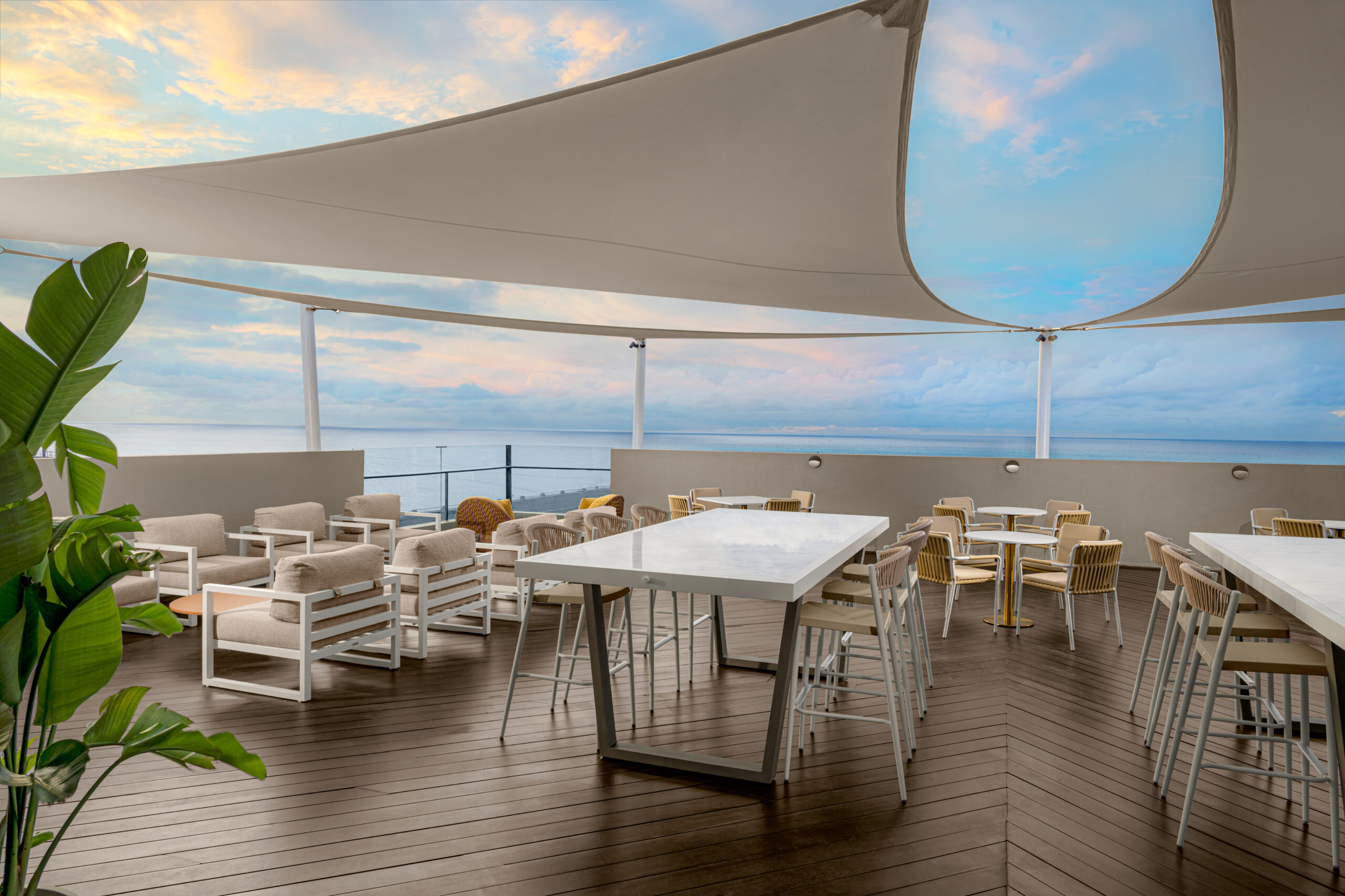 A modern outdoor terrace with white tables and chairs, wooden flooring, large shade sails overhead, and an ocean view in the background under a partly cloudy sky. A leafy green plant decorates the left side.