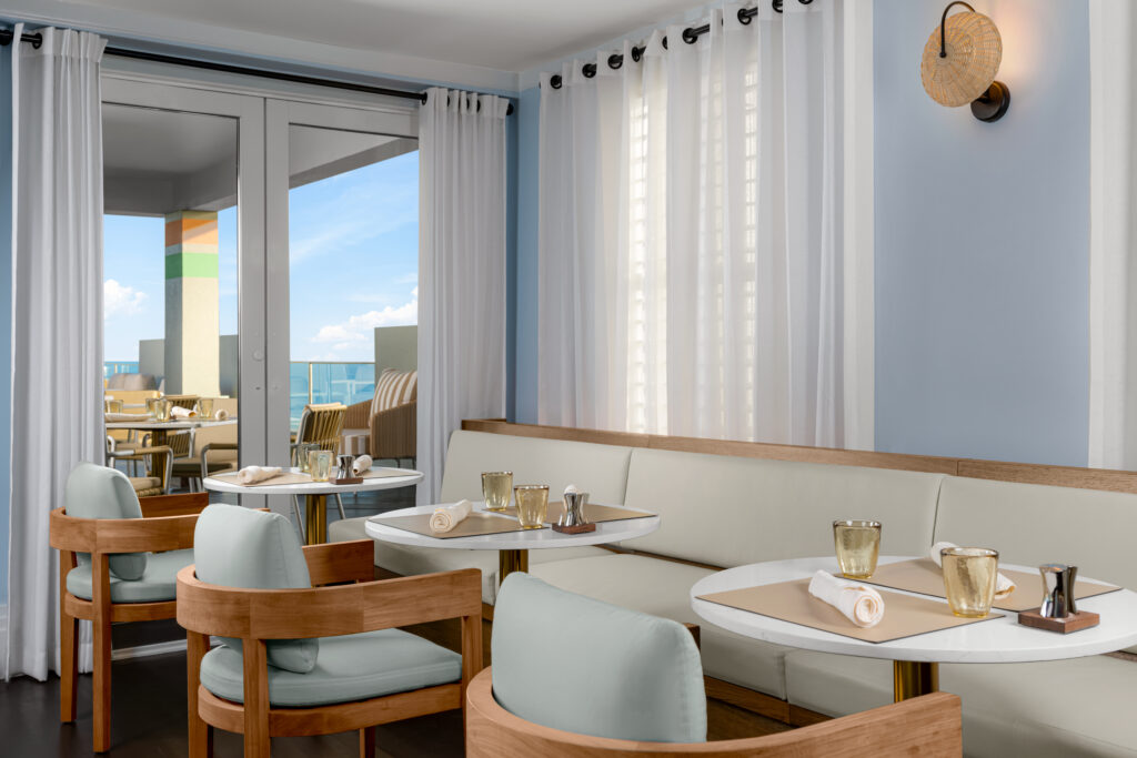 A modern, elegant restaurant with light blue walls, white curtains, and wooden furniture. Tables are set with glassware and napkins. Large windows offer a view of a sunny outdoor patio and blue sky.