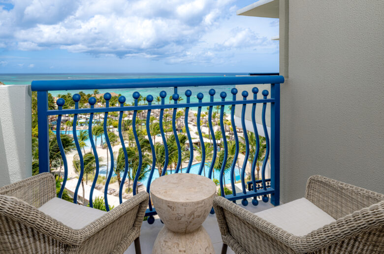 Two wicker chairs and a round stone table on a balcony with a blue railing, overlooking tropical pools, palm trees, and the turquoise ocean under a partly cloudy sky.