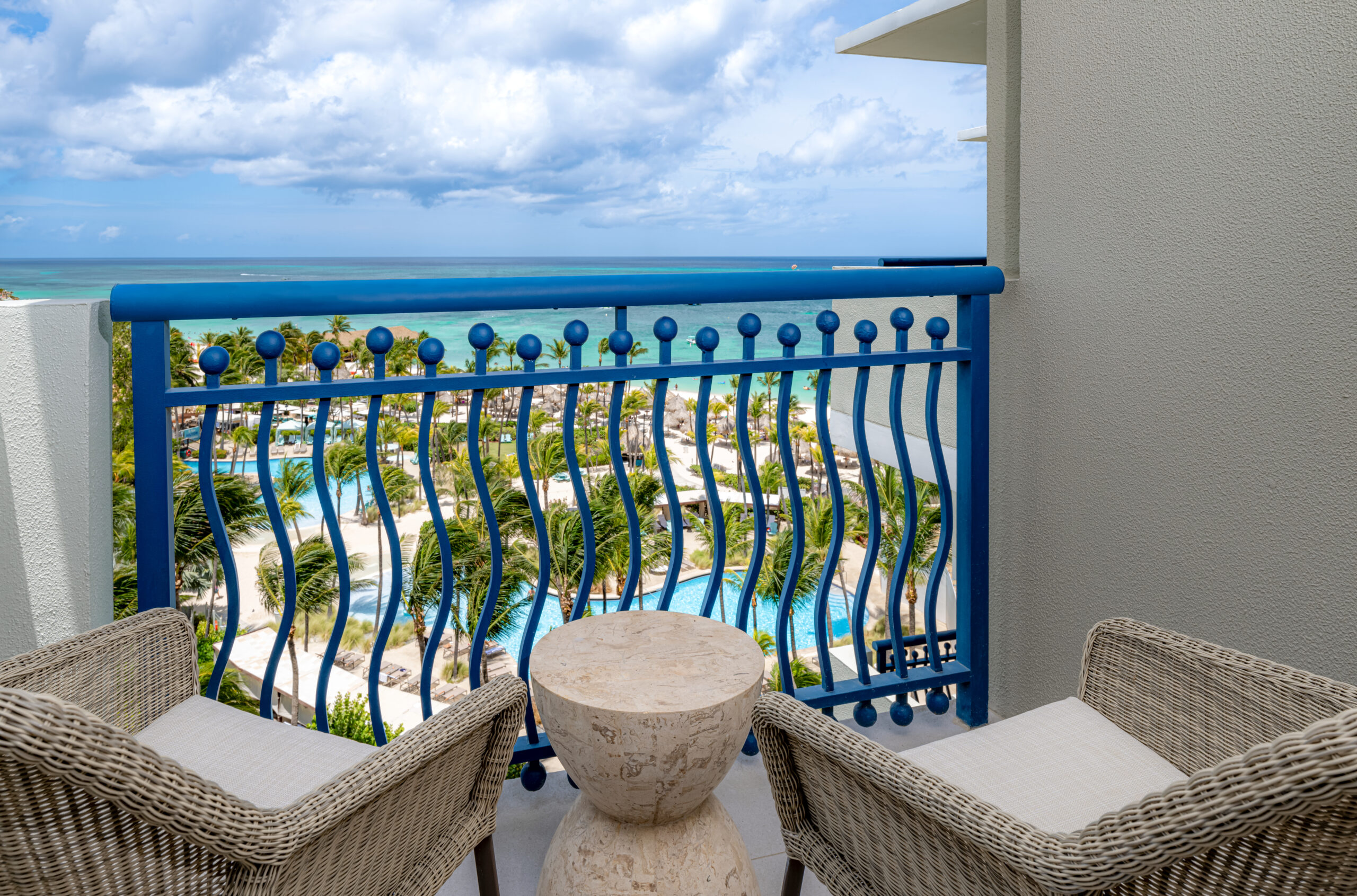 Two wicker chairs and a round stone table on a balcony with a blue railing, overlooking tropical pools, palm trees, and the turquoise ocean under a partly cloudy sky.