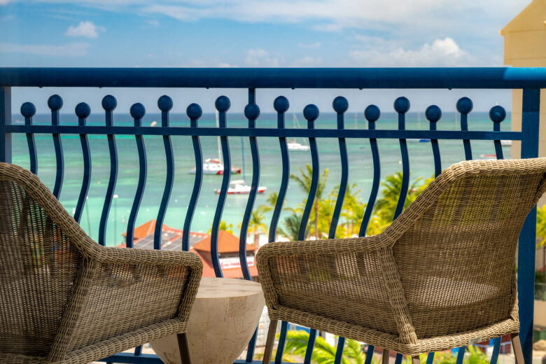 Two wicker chairs and a small round table sit on a balcony overlooking a turquoise ocean with boats, palm trees, and rooftops visible below. Blue sky with some clouds is in the background.