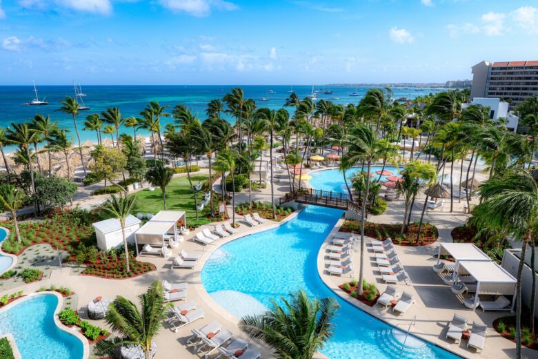 Aerial view of a tropical resort with palm trees, multiple pools, lounge chairs, cabanas, and a sandy beach overlooking a calm blue ocean with boats and a partly cloudy sky.