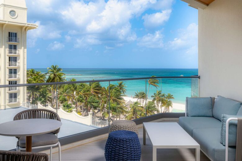A balcony with modern seating overlooks palm trees, a sandy beach, and turquoise ocean under a blue sky with scattered clouds.