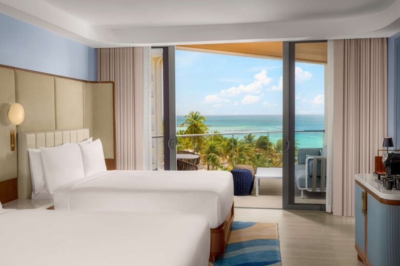 A hotel room with two beds, a balcony, and a view of palm trees and the ocean under a blue sky. The room has modern decor, a coffee maker, and neutral tones.
