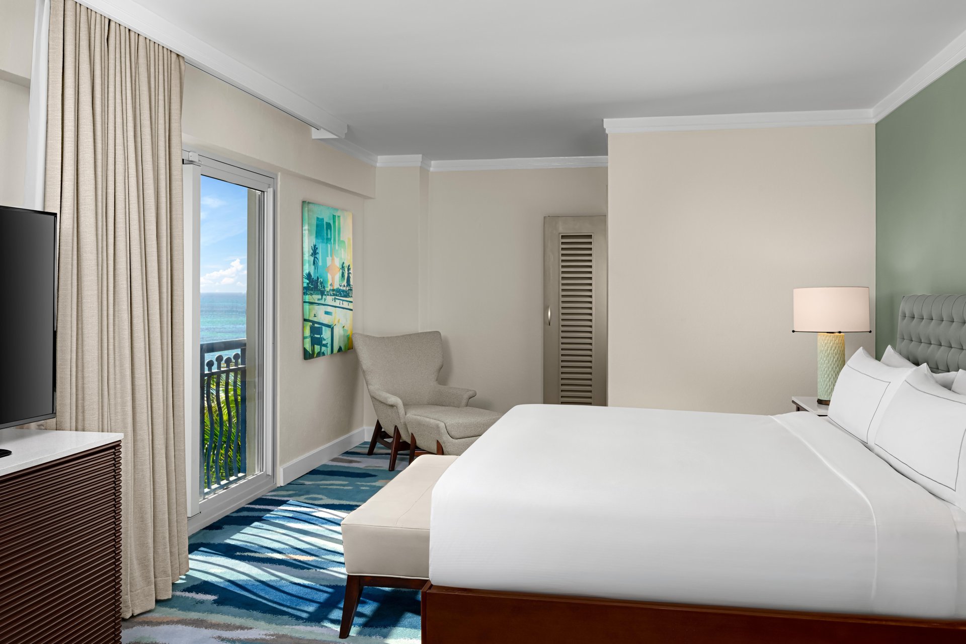A bright hotel room with a large bed, armchair, TV, lamp, and a balcony door that opens to an ocean view. The room has modern decor, light-colored walls, and a blue-patterned carpet.