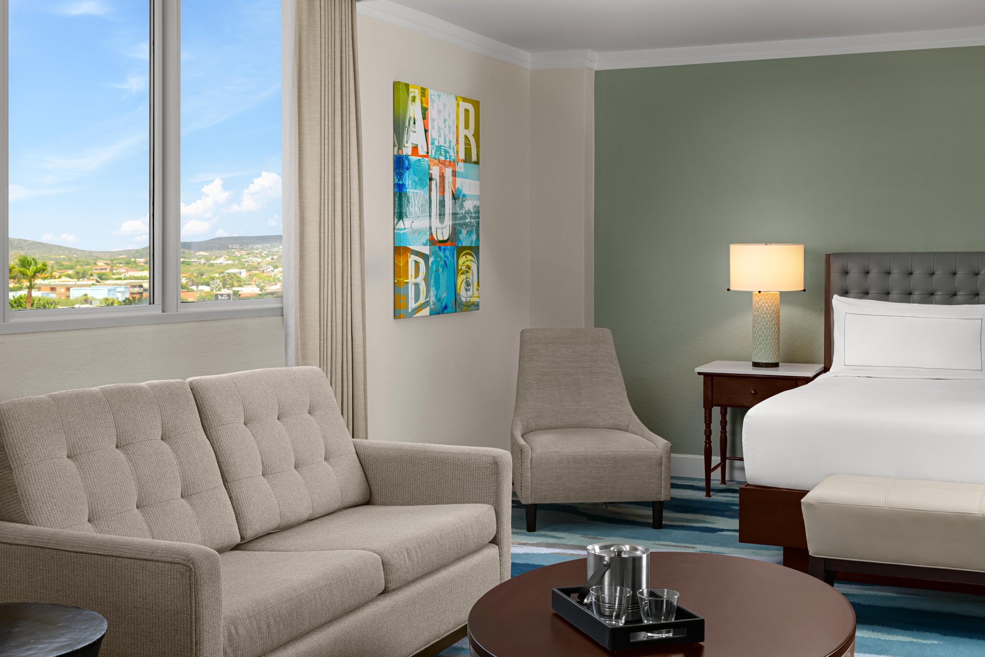 A modern hotel room with a beige sofa, armchair, round coffee table, and a bed with white linens. A lamp sits on a nightstand, and a window offers a view of a bright, hilly landscape outside.