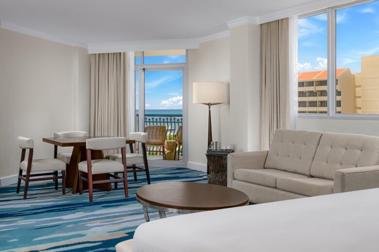 A bright hotel suite with a beige sofa, round coffee table, dining table with four chairs, large windows, and a balcony offering views of buildings and a partly cloudy sky. Blue patterned carpet covers the floor.