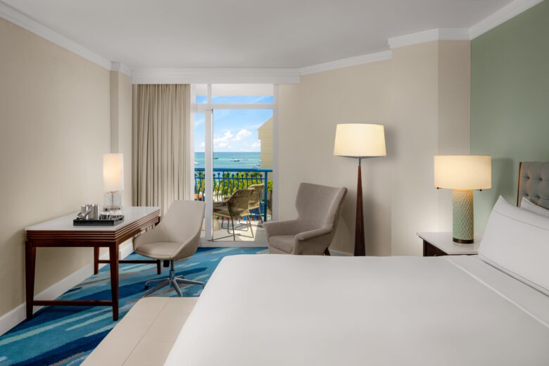 A bright hotel room with a bed, desk, chair, armchair, two lamps, and a large window leading to a balcony with an ocean view. Neutral walls and blue accents create a calm, inviting atmosphere.