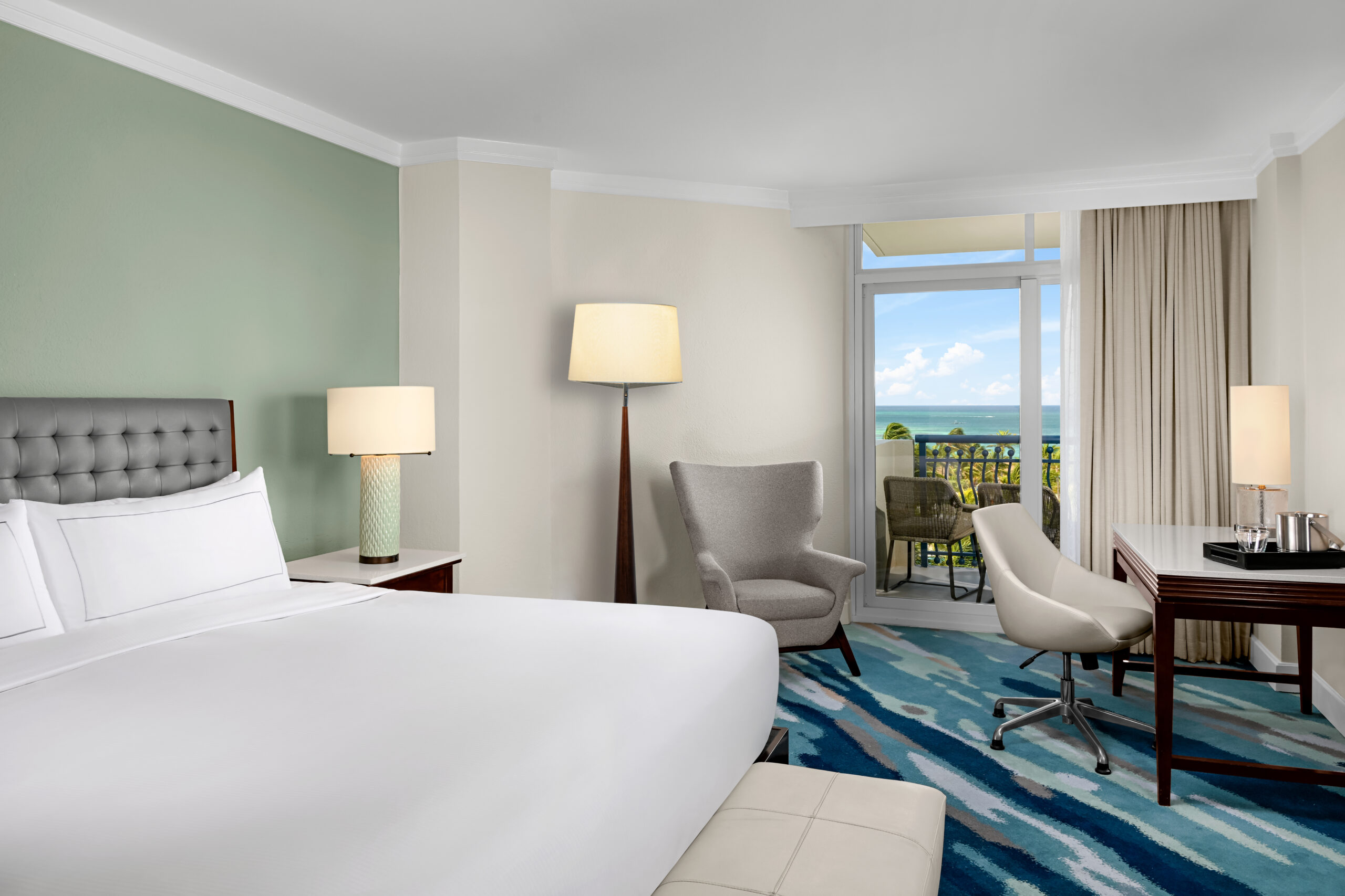 A modern hotel room with a large bed, armchair, desk, and lamps. A balcony with chairs overlooks the ocean, visible through glass doors, and the room features blue patterned carpet and soft green and white walls.