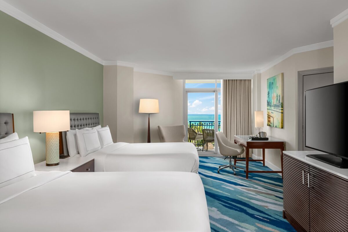 Modern hotel room with two double beds, lamps, a TV, desk and chair, armchair by a lamp, and a balcony overlooking the ocean. Artwork decorates the wall, and the room features blue patterned carpet.