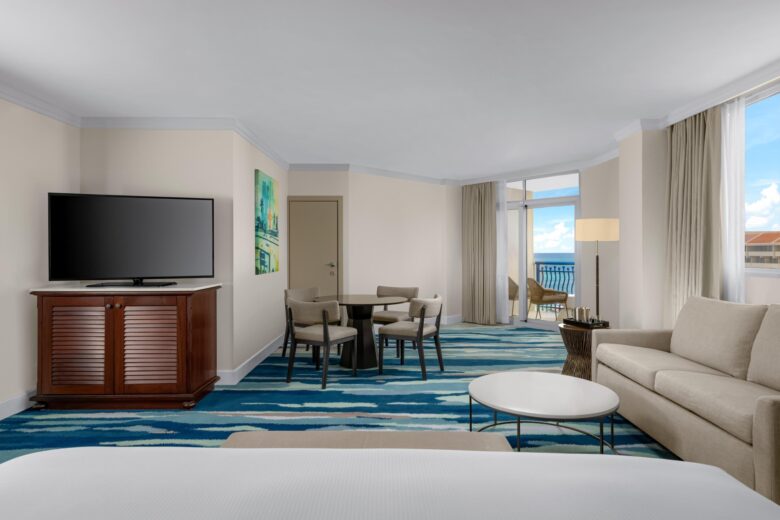 A spacious hotel room with a bed in the foreground, a beige sofa, a round coffee table, a dining table with four chairs, a flat-screen TV on a wooden stand, large windows, and a balcony with an ocean view.