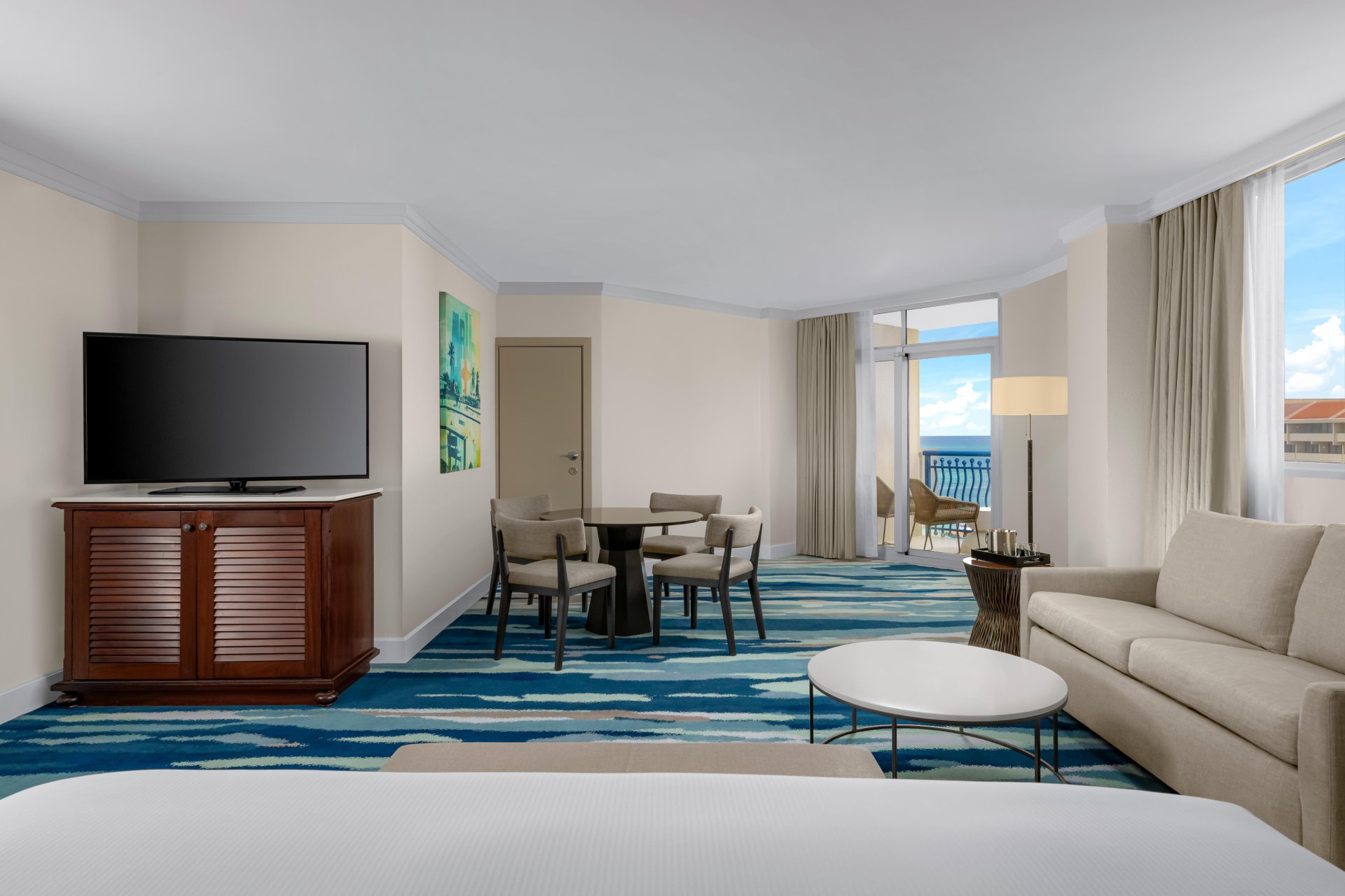 A spacious hotel room with a bed in the foreground, a beige sofa, a round coffee table, a dining table with four chairs, a flat-screen TV on a wooden stand, large windows, and a balcony with an ocean view.