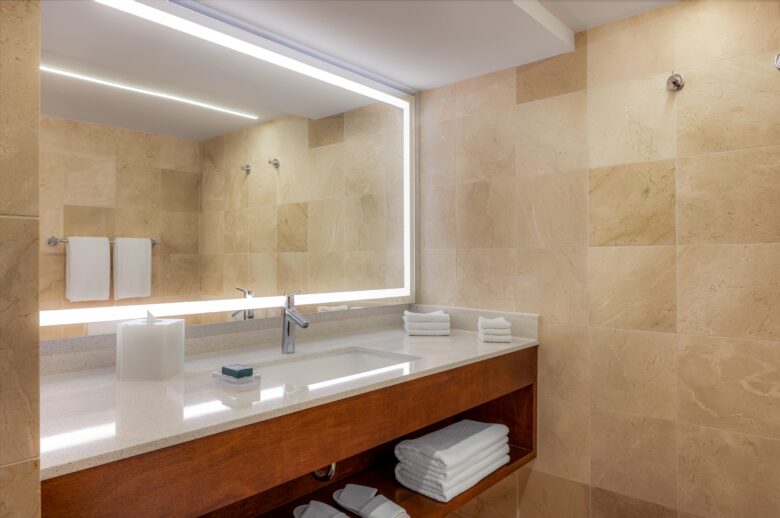 Modern bathroom with beige tiled walls, a large illuminated mirror, a white countertop with a built-in sink, a faucet, a tissue box, soap, neatly folded towels on and under the counter, and towel racks on the wall.