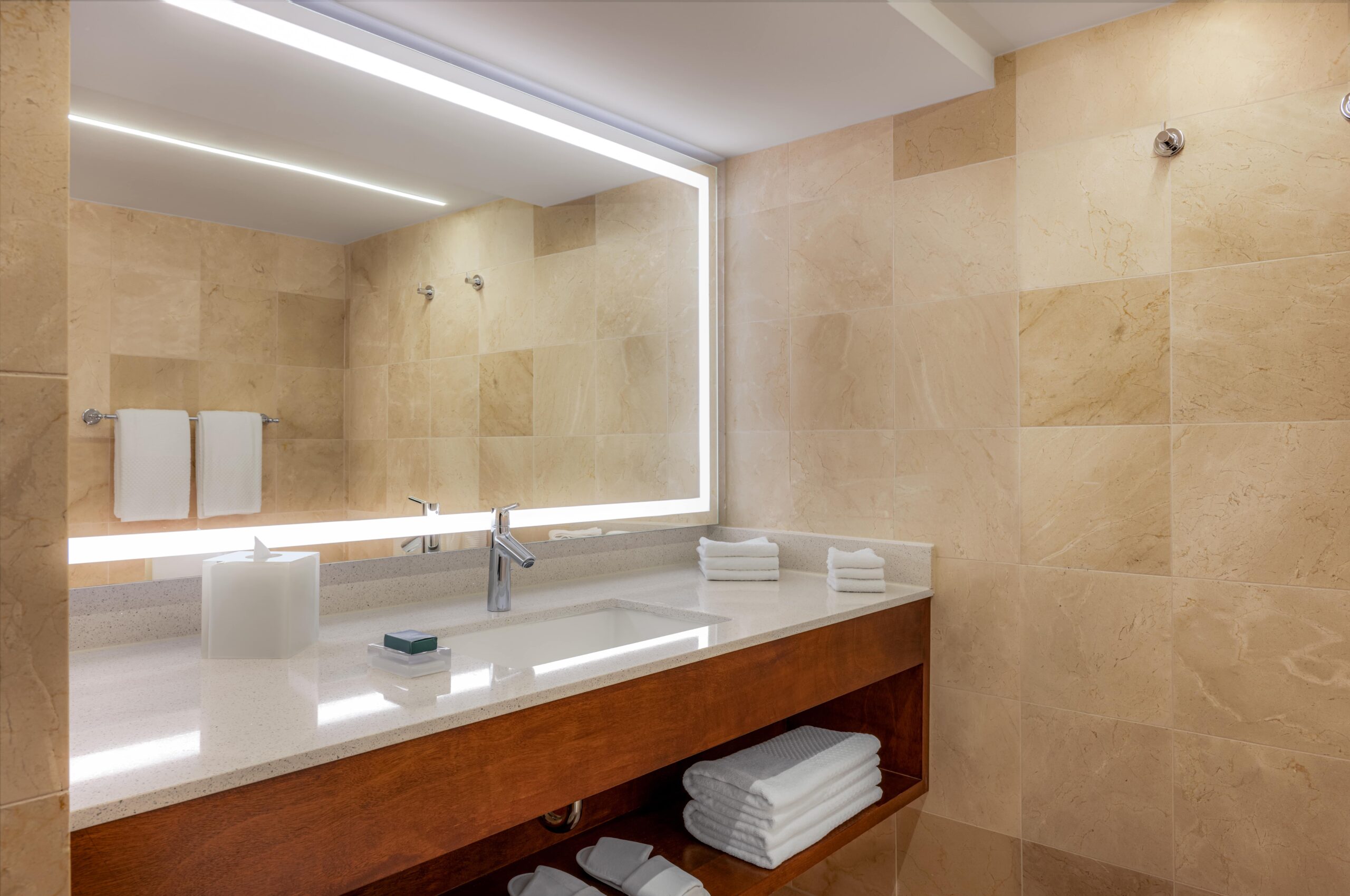 Modern bathroom with beige tiled walls, a large illuminated mirror, a white countertop with a built-in sink, a faucet, a tissue box, soap, neatly folded towels on and under the counter, and towel racks on the wall.