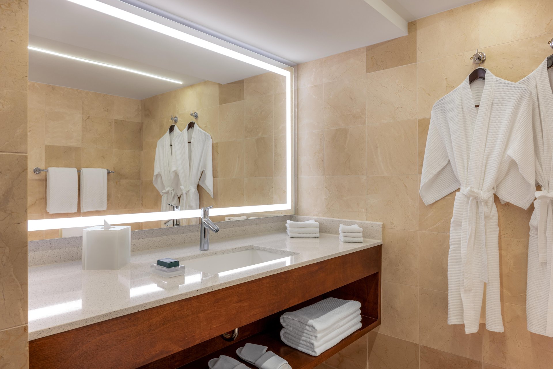 Modern bathroom with a large illuminated mirror, towels, and two white bathrobes hanging on beige tiled walls, a countertop with a sink, soap, and neatly folded towels stored beneath.