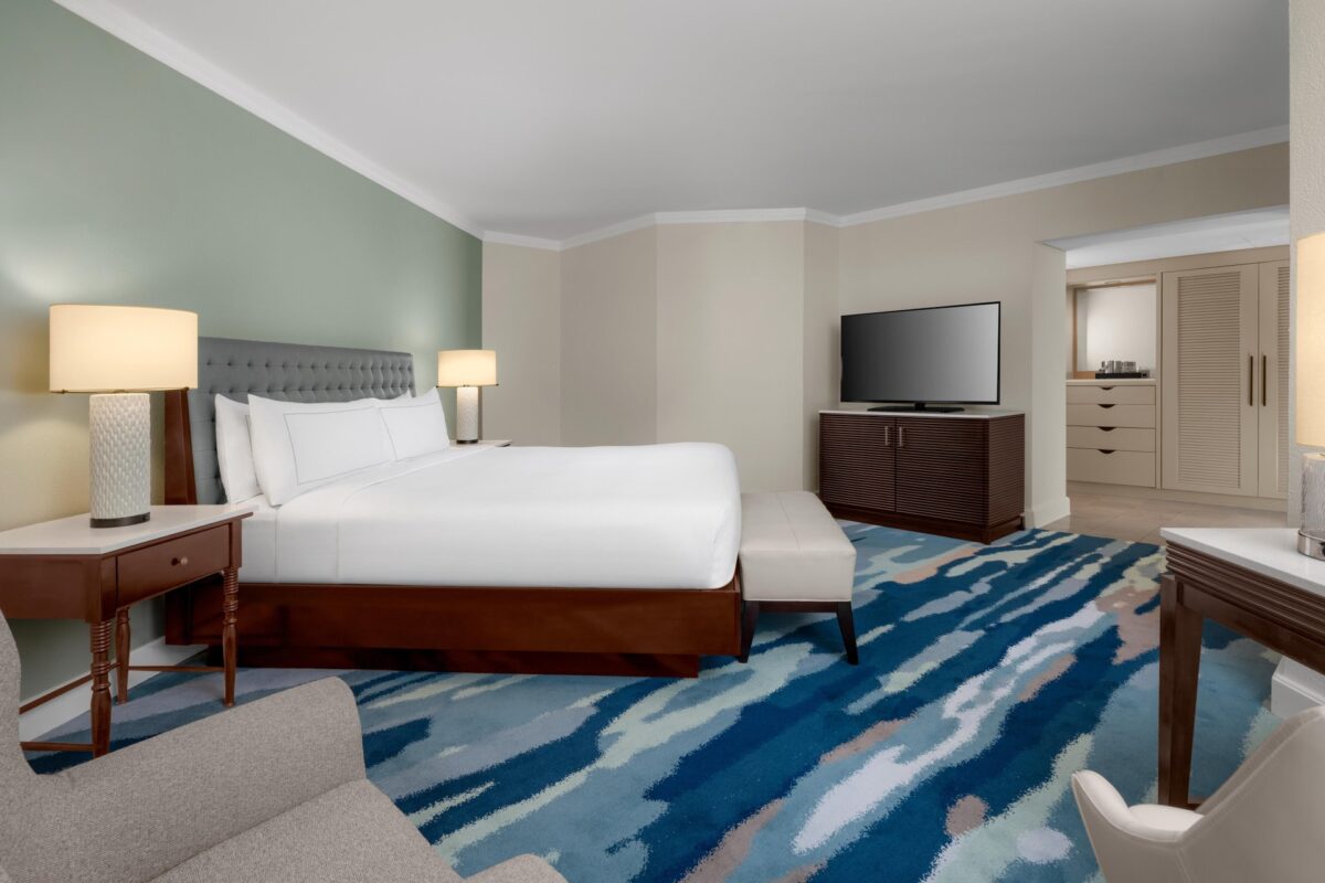 A modern hotel room with a king-sized bed, two bedside tables with lamps, a TV on a cabinet, a patterned blue carpet, an armchair, and a desk with a chair. Light-colored walls and a closet are also visible.