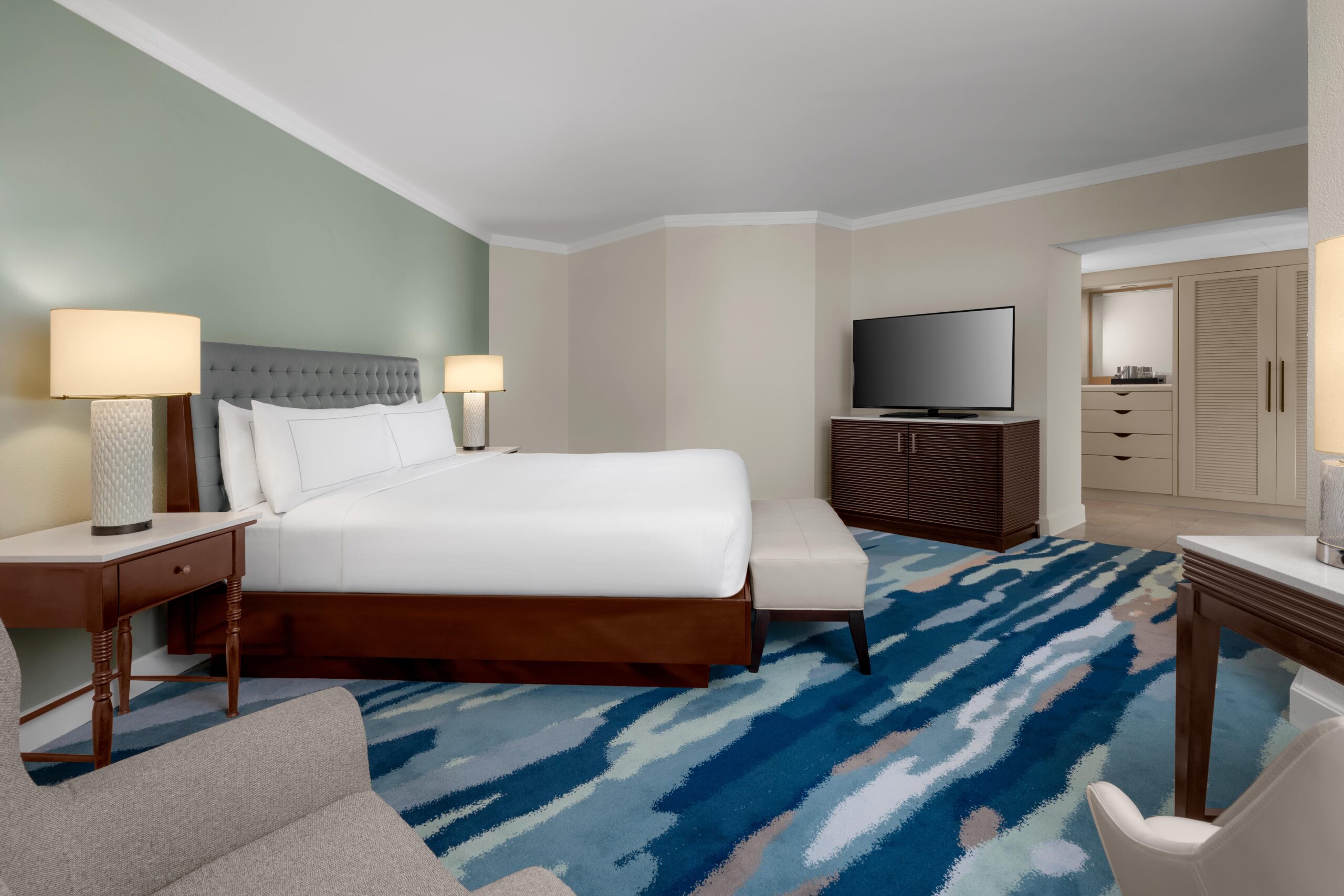 Modern hotel room with a king-size bed, nightstands with lamps, a flat-screen TV on a dresser, cushioned bench, desk, and a patterned blue area rug. Room has neutral walls and soft lighting.