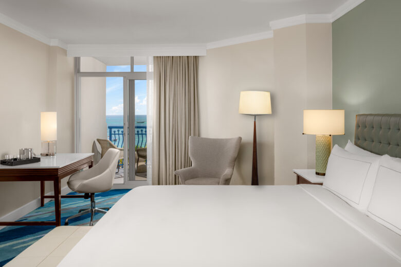 A modern hotel room with a neatly made bed, a desk and chair, an armchair with a floor lamp, and a balcony with an ocean view through large glass doors. The decor is light and calming.