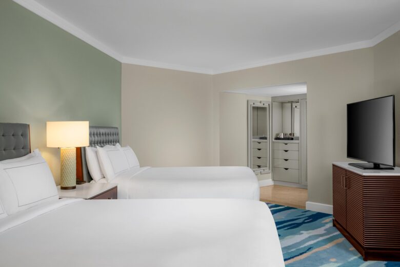 A hotel room with two double beds, a bedside table with a lamp, a flat-screen TV on a wooden dresser, and an open doorway leading to a bathroom with a large mirror. The room has light-colored walls and a patterned rug.