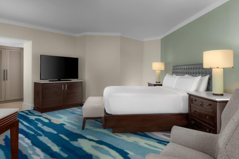 Modern hotel room with a king-sized bed, two bedside tables with lamps, a flat-screen TV on a dresser, an armchair, and a blue patterned carpet. Walls are light beige and green.