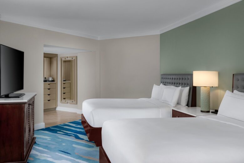 A hotel room with two large beds, a flat-screen TV on a wooden cabinet, a nightstand with a lamp, an abstract blue rug, and a closet with a mirror and drawers in the background.