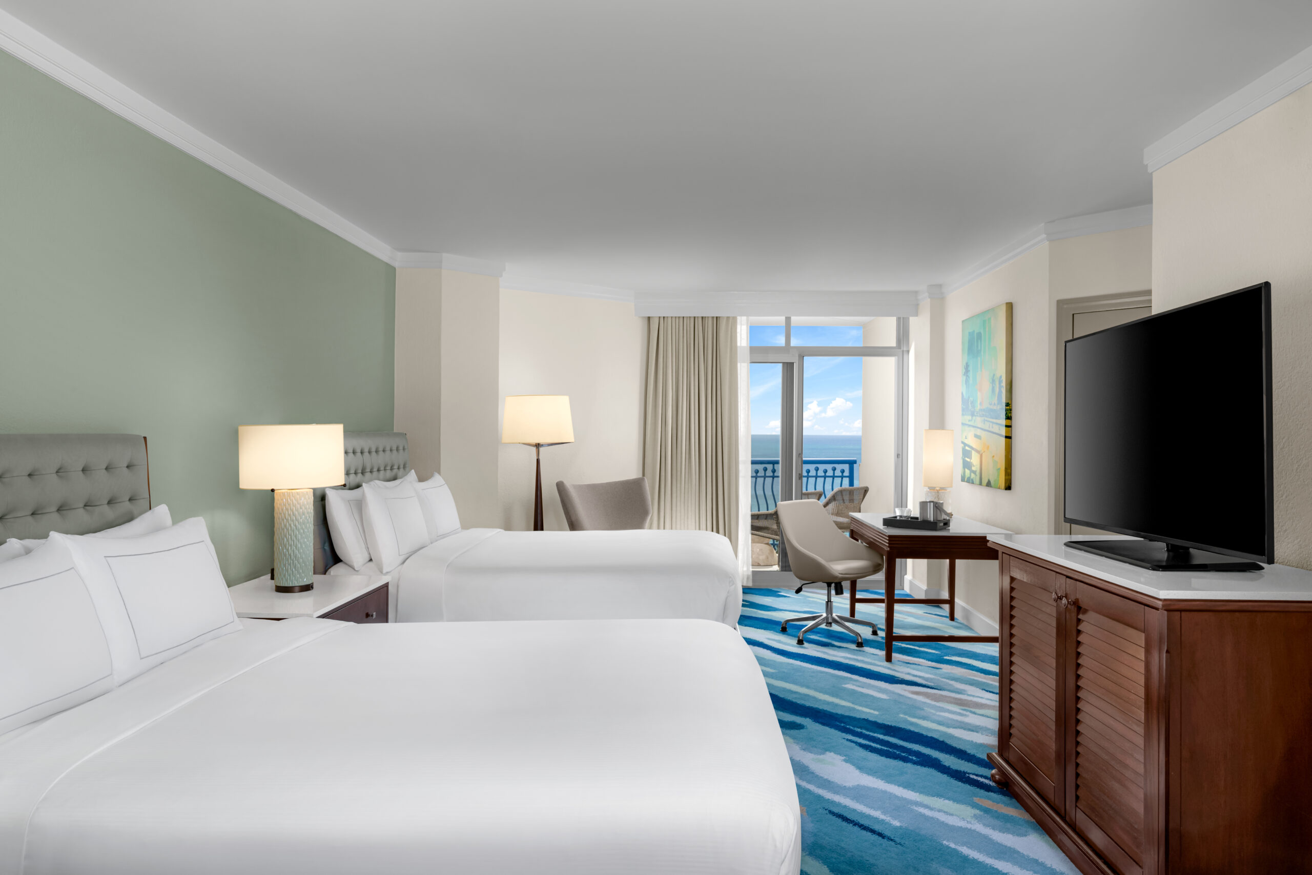 A hotel room with two double beds, a large TV on a wooden dresser, a work desk and chair by a window, modern lamps, and a balcony with an ocean view. The decor is light and contemporary.
